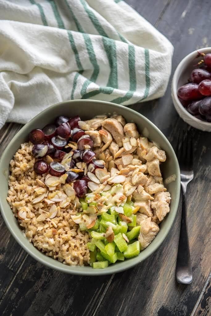 A bowl of healthy chicken salad brown rice bowl with grapes and celery with a napkin and a fork and bowl of grapes