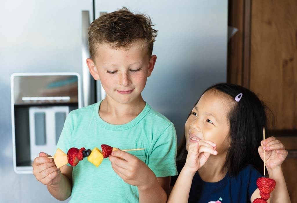 Two children smiling and making fruit kabobs
