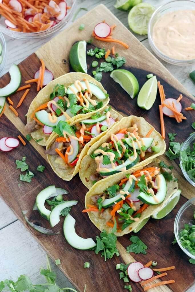 Overhead view of Banh Mi Tacos on a wooden platter with veggies and limes