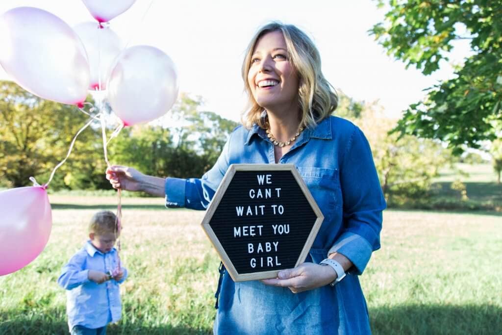 A Mom holding pink balloons and a sign with an baby girl announcement