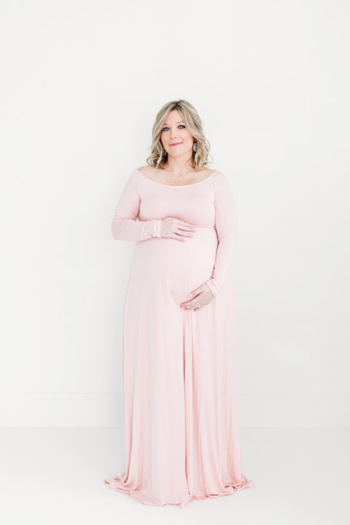 A pregnant woman in a pink dress holding her belly and smiling during maternity photos