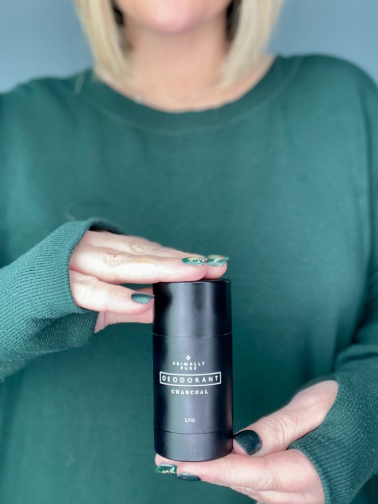 a woman with blonde hair and a green shirt holding a bottle of primally pure deodorant