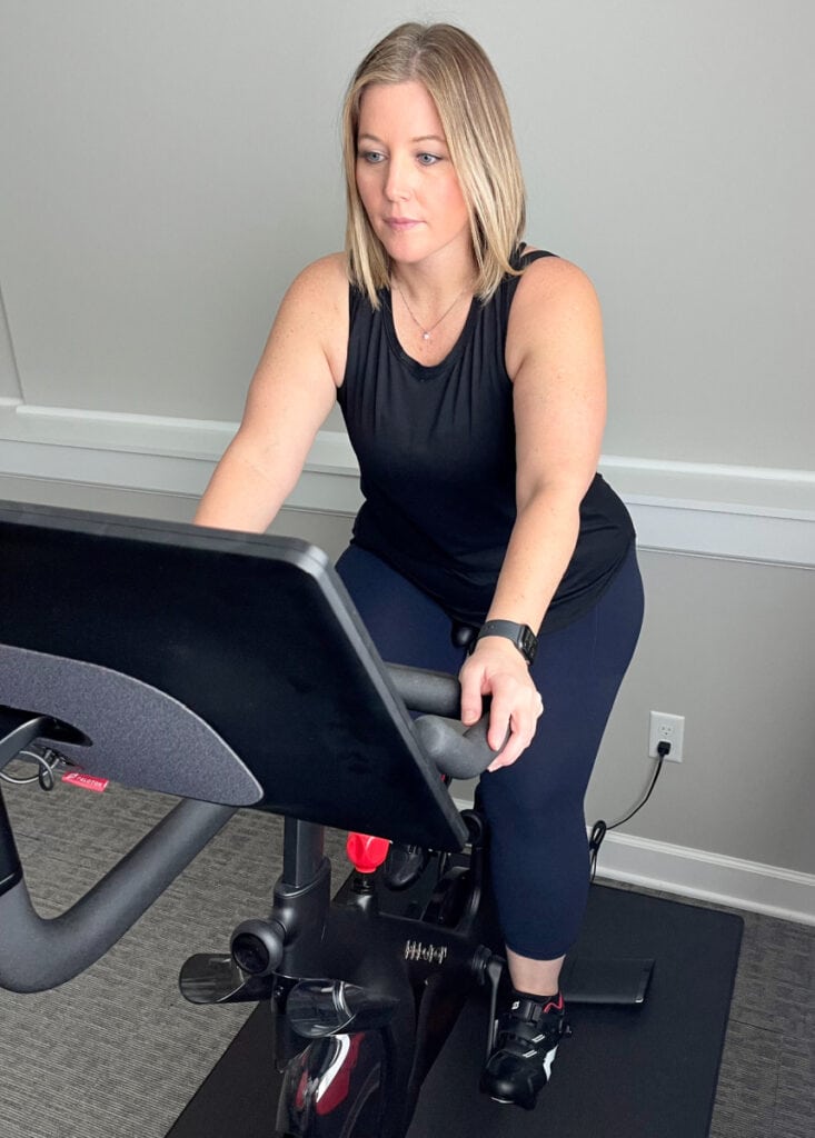 a blonde woman sitting on a peloton bike  with a black shirt on and blue leggings