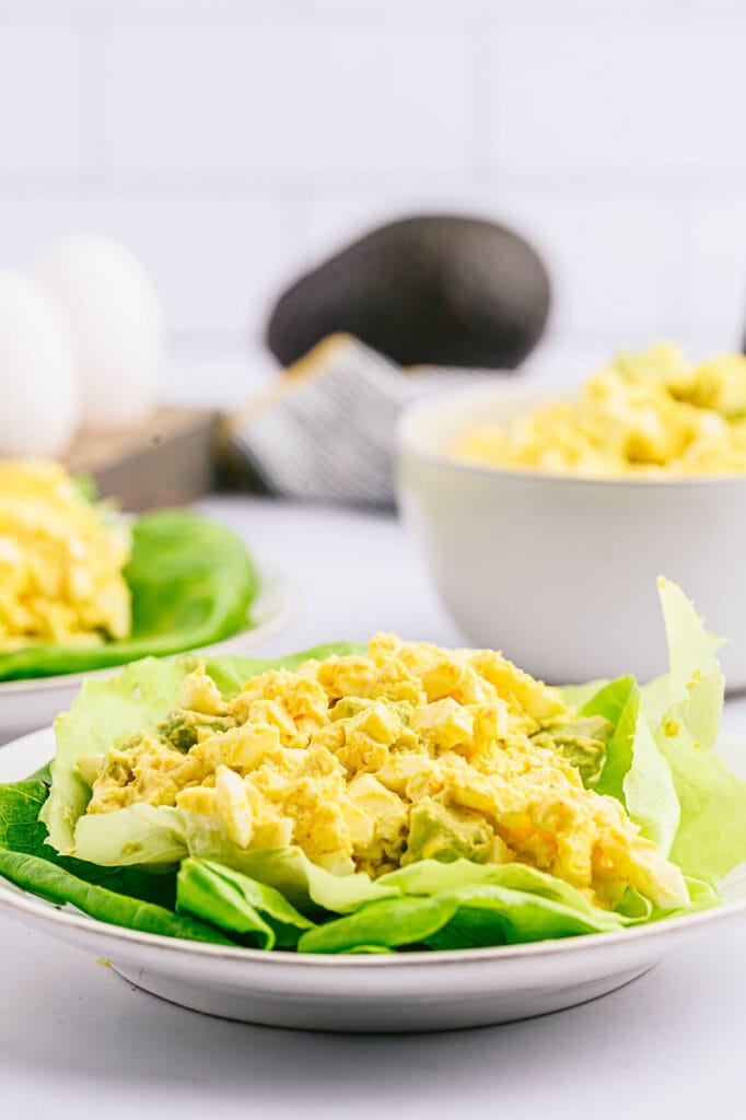 Avocado egg salad on a bed of lettuce and then two bowls of the salad in the background and an avocado in the background