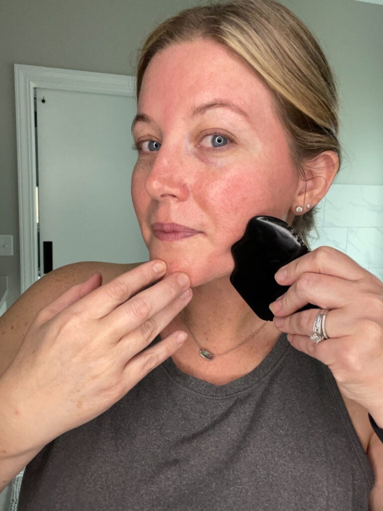 a blonde haired woman with a grey shirt on using a gua sha stone on her jaw
